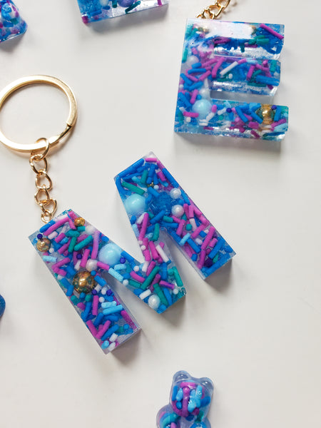 resin alphabet letter keychains made with real cake sprinkles that are blue, purple and green attached with gold keychain hardware.