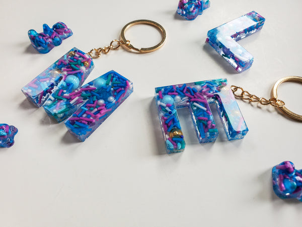 resin alphabet letter keychains made with real cake sprinkles that are blue, purple and green attached with gold keychain hardware.