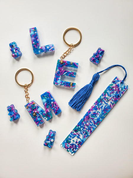 resin alphabet letter keychains made with real cake sprinkles that are blue, purple and green attached with gold keychain hardware.