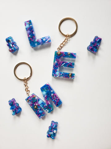resin alphabet letter keychains made with real cake sprinkles that are blue, purple and green attached with gold keychain hardware.