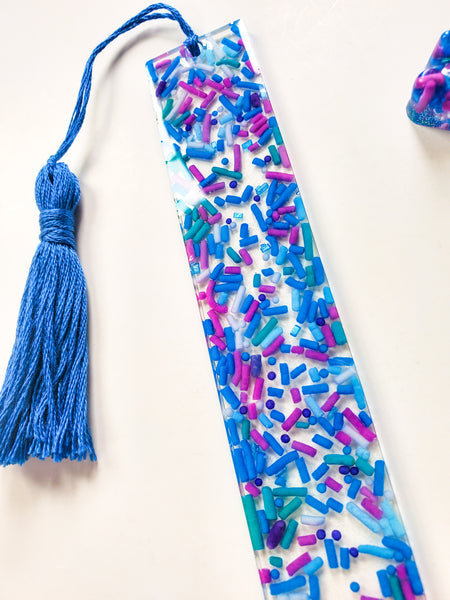 a resin bookmark made with real cake sprinkles that are blue, purple and green with a handmade tassel made with matching blue cotton