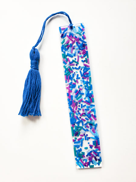 a resin bookmark made with real cake sprinkles that are blue, purple and green with a handmade tassel made with matching blue cotton